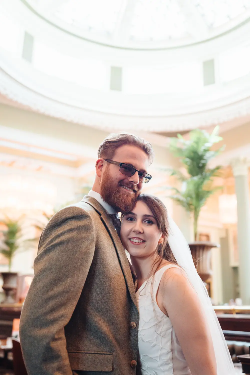 Sarah and James posing for a romantic couple portrait inside The Cosy Club Norwich, the venue's beautiful domed rotunda ceiling and tall plants framing them from above