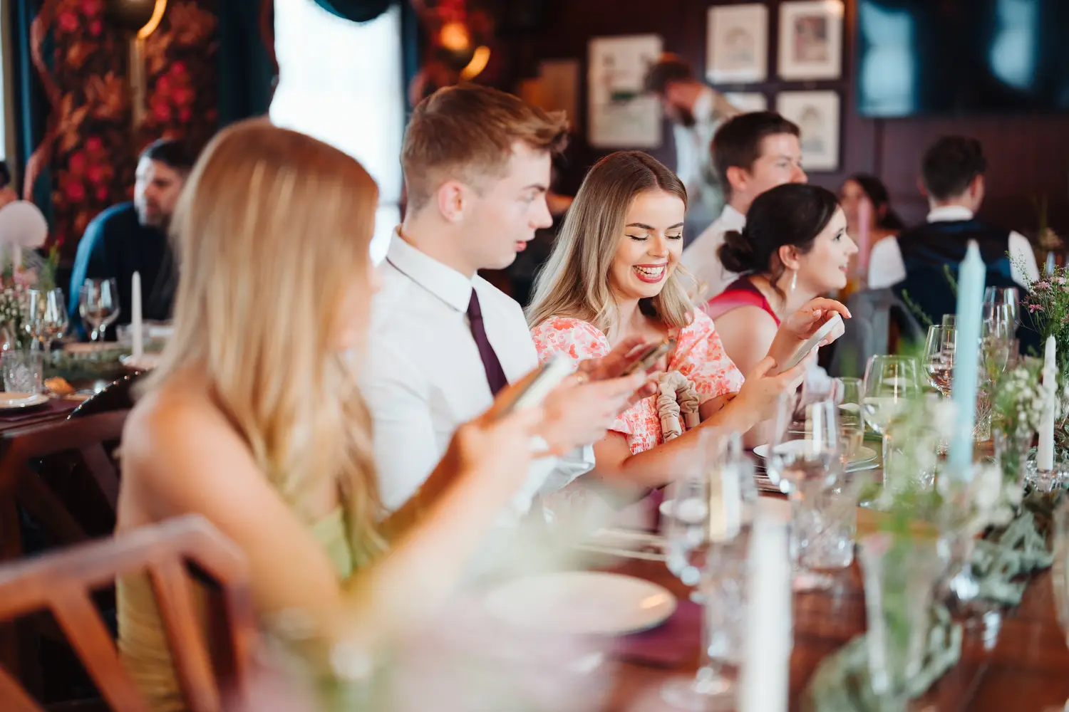Young wedding guests smiling and checking their phones at the dinner table during the reception at The Cosy Club Norwich, candlelit table setting around them