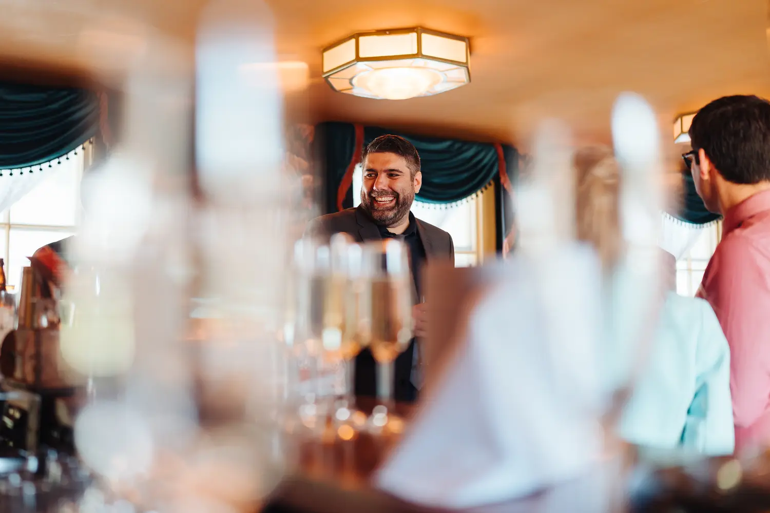 Male wedding guest laughing with another guest through sparkling champagne glasses at The Cosy Club Norwich wedding reception, bokeh lights twinkling behind them