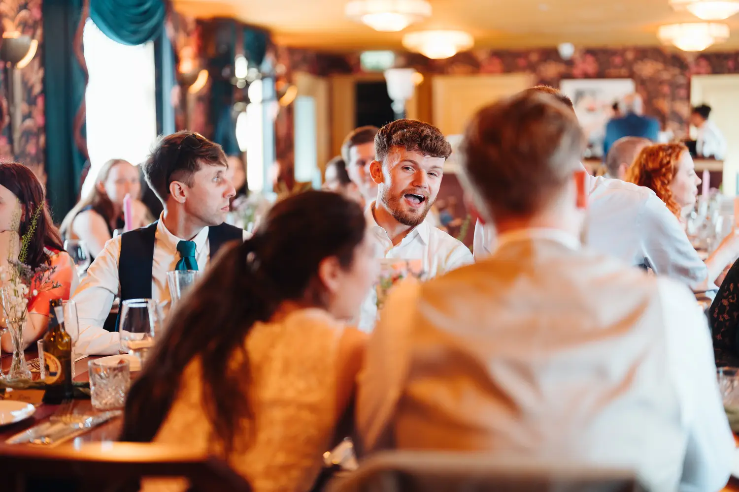 Animated male wedding guest laughing and talking at the dinner table during Sarah and James's reception at The Cosy Club Norwich, other guests visible in the background