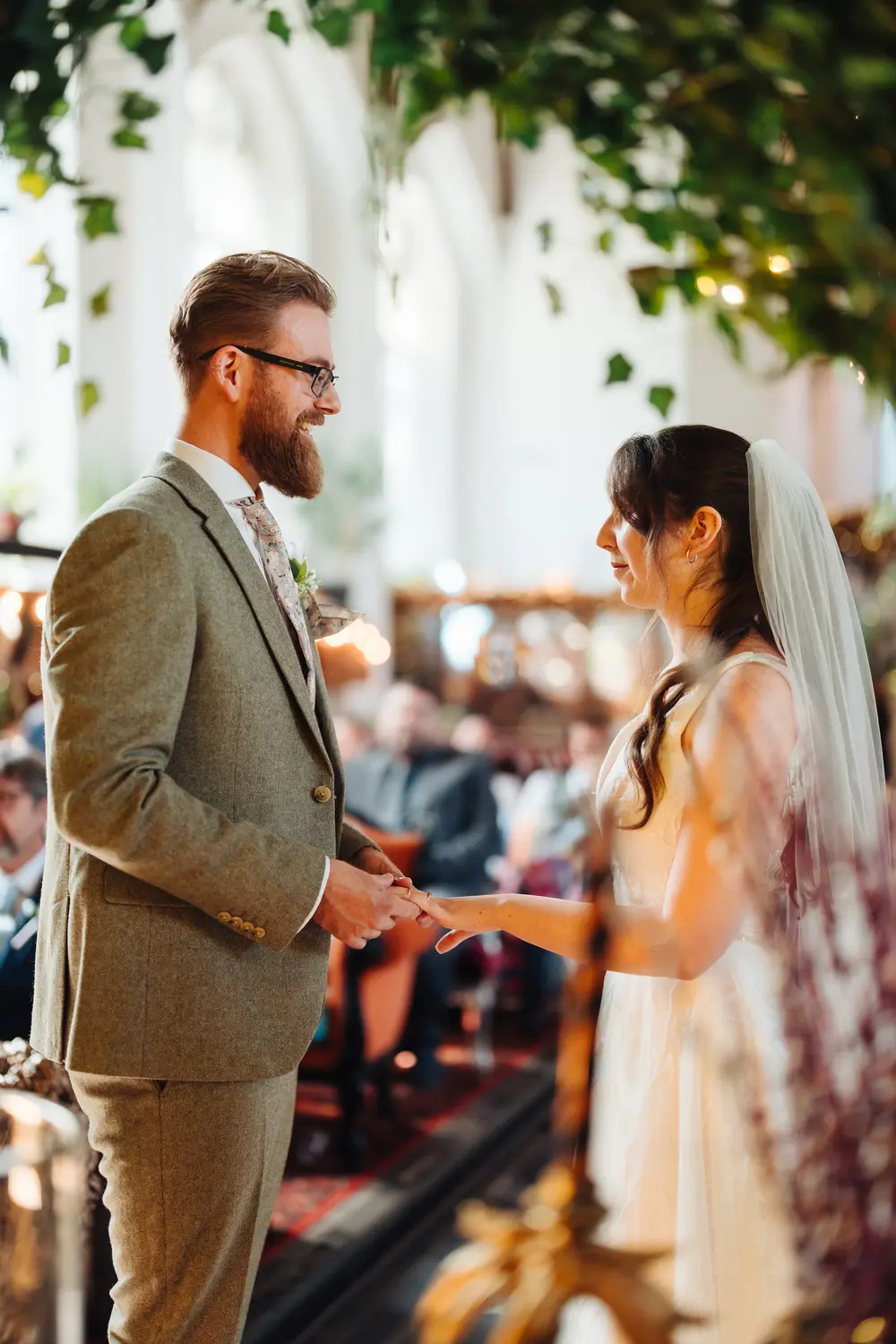 James placing the wedding ring on Sarah's finger during their ceremony at The Flint Rooms Norwich, ivy trailing overhead from the gothic arched ceiling