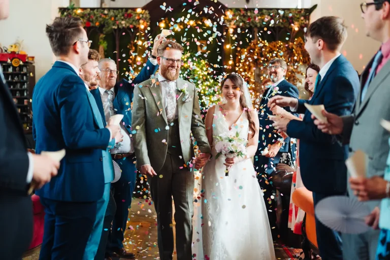 Sarah and James showered in colourful confetti as they walk the long aisle at The Flint Rooms Norwich, guests cheering on both sides in the eclectic venue