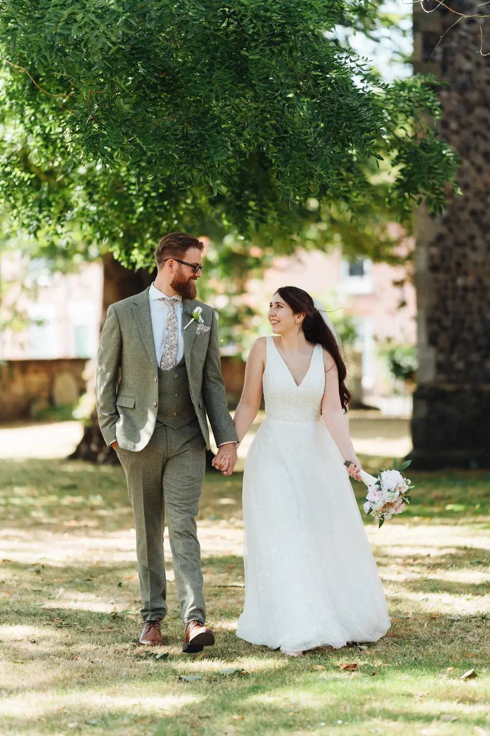 Sarah and James walking hand in hand through the sun-dappled gardens near The Flint Rooms Norwich, smiling at each other with Sarah holding her bouquet