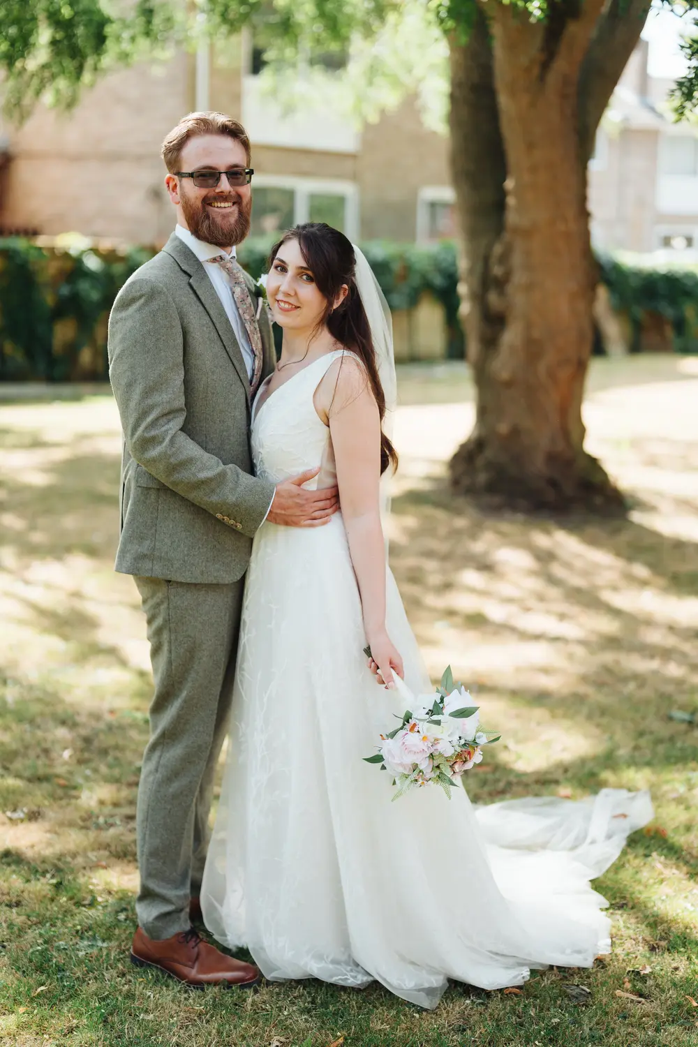 Sarah and James posing together in the gardens outside The Flint Rooms Norwich, James holding Sarah from behind with a warm smile on their wedding day