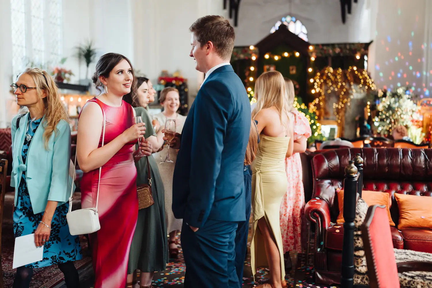 Wedding guests mingling in colourful outfits with drinks inside The Flint Rooms Norwich after the ceremony, fairy lights and vintage decor filling the room