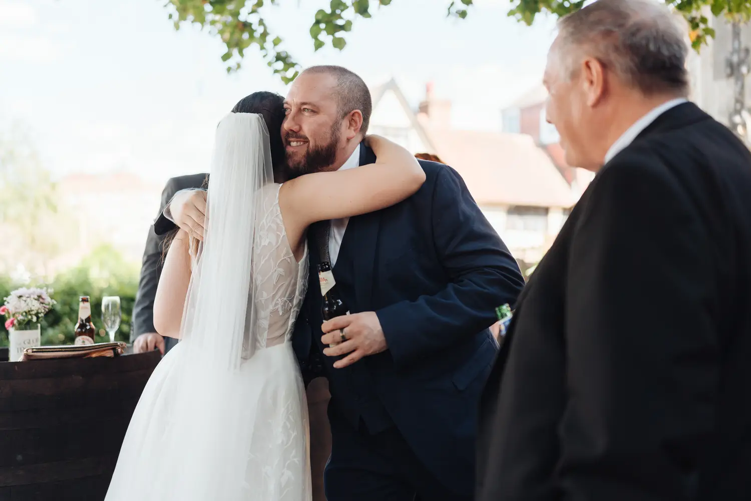 Sarah hugging and laughing with a male guest in a blue suit outside The Flint Rooms Norwich, trees and brick buildings visible in the warm afternoon light