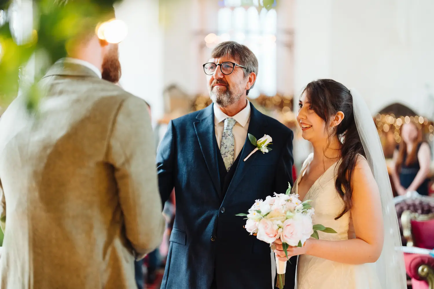 Sarah smiling at James at the altar as her father stands proudly beside her holding her bouquet of white and pink roses at The Flint Rooms Norwich