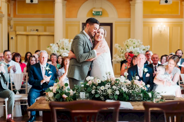 Exuberant newlywed couple sharing a hug during their reception among many seated friends and family.
