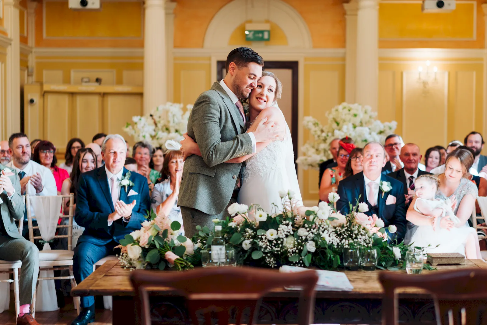 Exuberant newlywed couple sharing a hug during their reception among many seated friends and family.